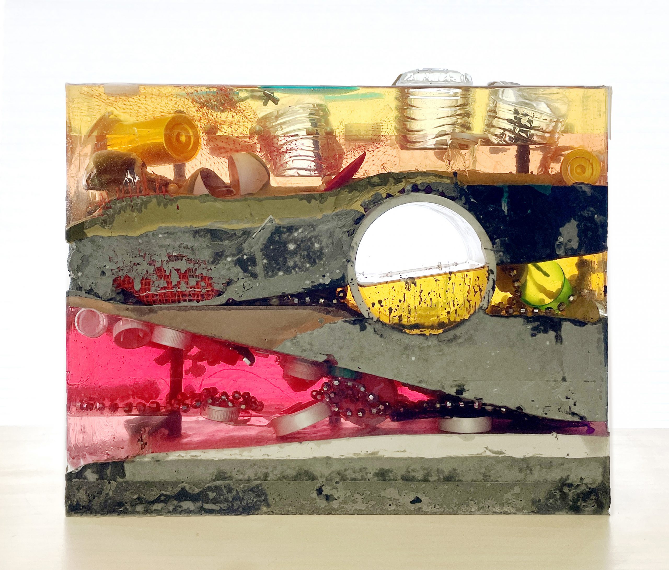 Transparent resin block with layered colors encasing everyday objects—bottle caps, toys, and fragments—arranged like a cross-section of buried debris.