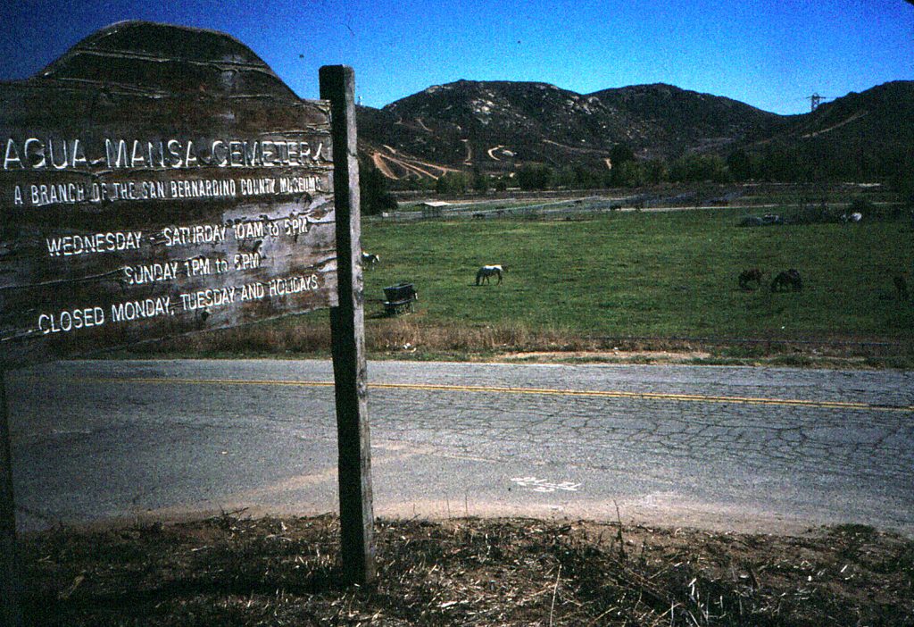 A paved road is seen in the center of the landscape photos with cattle grazing in the background and an aging Agua Mansa Cemetery wood sign is seen in the far left forefront in dirt.