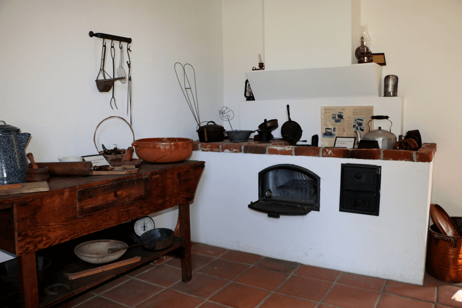 An 1800s kitchn with brick oven and wooden table to the left. Cast iron pans and metal tea kettle can be seen on the rustic adobe countertop/ Red clay tiles line the floor of this kitchen.