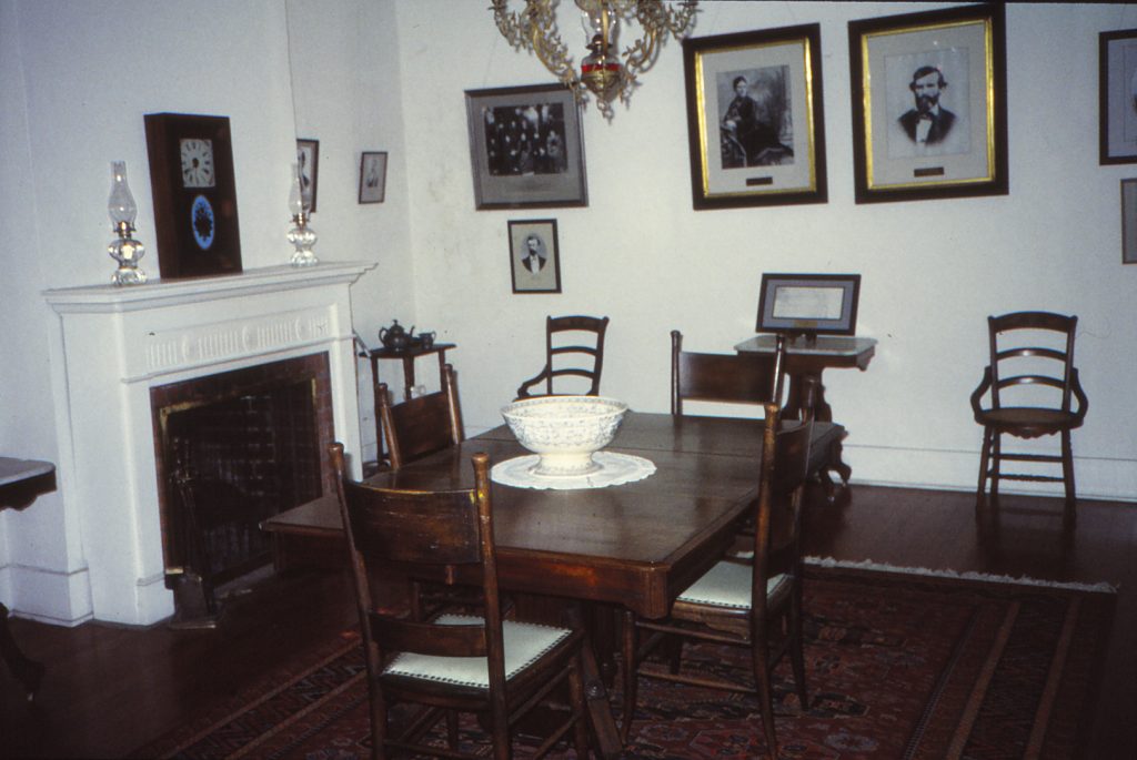 An interior shot of the Maria Merced Williams and John Rains' house dining room with fireplace showing on the left-hand side with white mantle and photos seen hanging in the background with a long wood table and chairs. The floor is also a dark hardwood original floor.