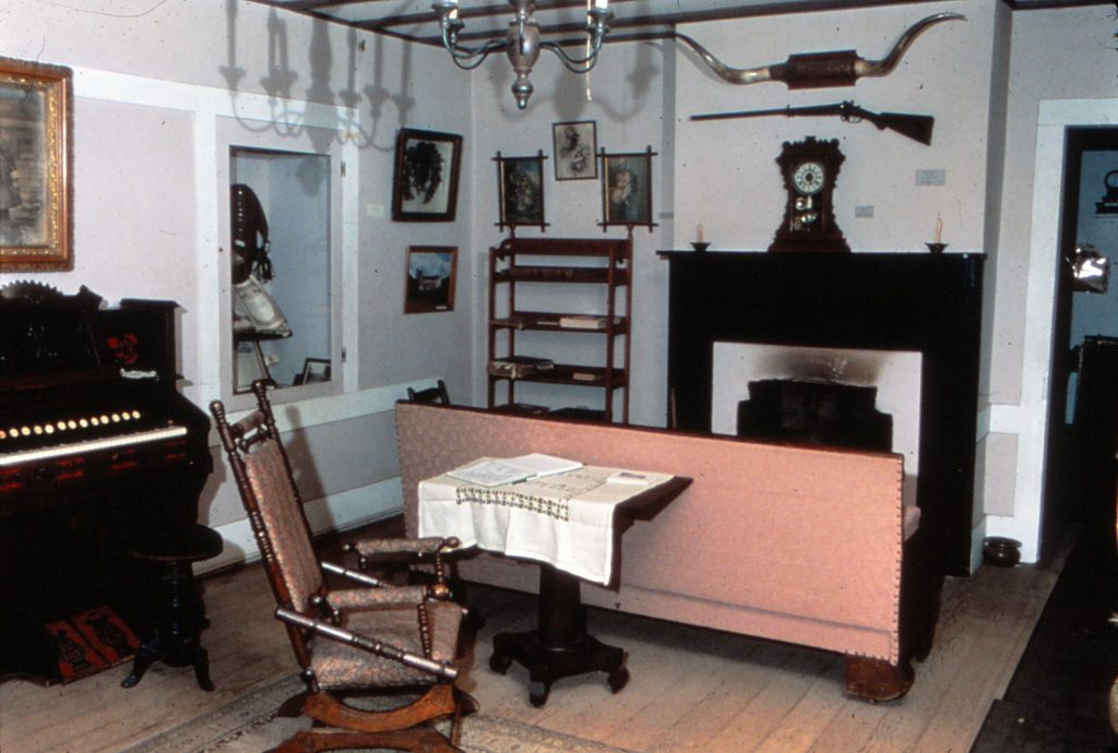 An inside view of the main room inside the Adobe. A mantle is seen in the background with a rifle hanging above. a rocking chair is seen in the left-hand side and back of a couch that faces the fireplace. Steer horns are seen hanging above the rifle and a candle chandelier is seen hanging from the ceiling.