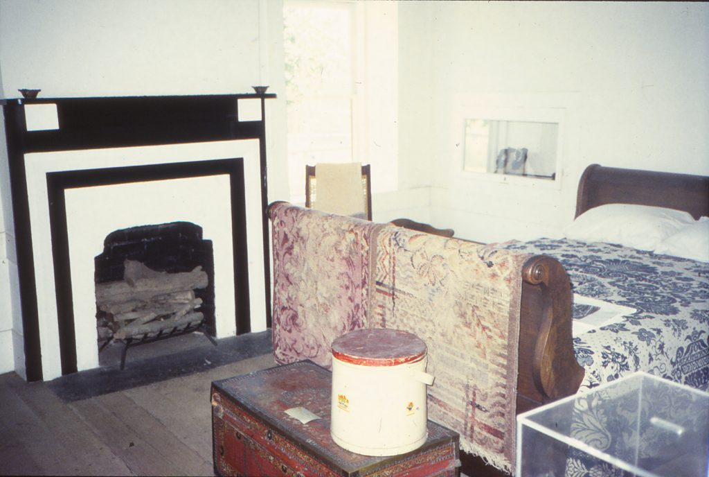 A view of a bedroom inside the Adobe. A fireplace is seen on the left-hand side and a sleigh bed is seen on the right-hand side of the room with a quilt blanket hanging from the footboard and a an old chest with tin canister is seen at the foot of the bed on the floor.