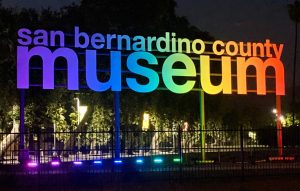 A night time picture of the San Bernardino County Museum sign outside the main museum in Redlands with color lights illuminating the sign in purple, blue light blue, yellow and red orange.
