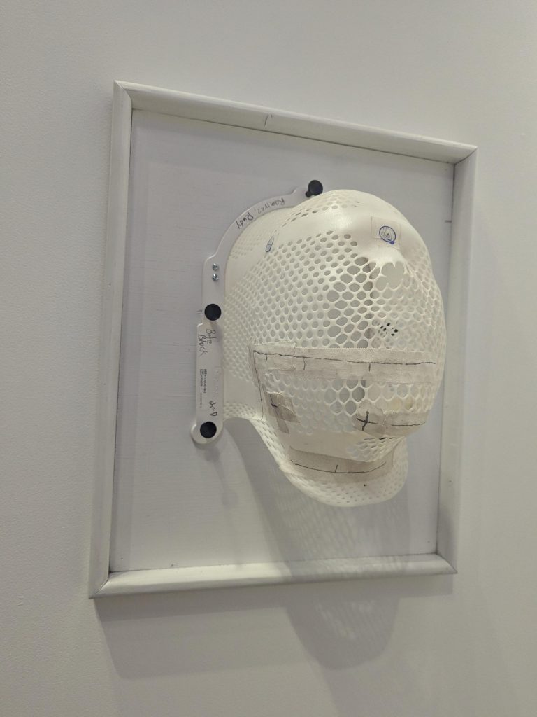 A front-left angled view of a white mesh-like face mask, molded to fit a head, affixed to a white board and frame. Visible handwritten notes and black fasteners surround the edges of the mask.
