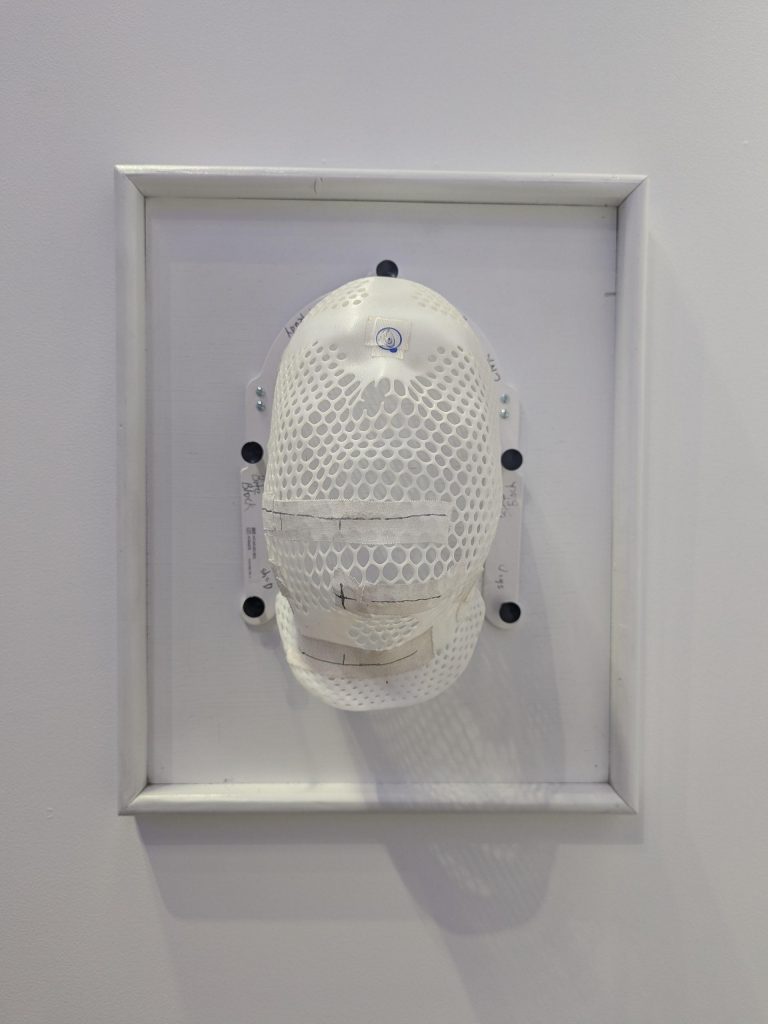 A centered, head-on view of a white perforated face mask attached to a white-framed board on a wall. The mask features pen markings and patches of medical tape, indicating it may have been used for medical or therapeutic purposes.