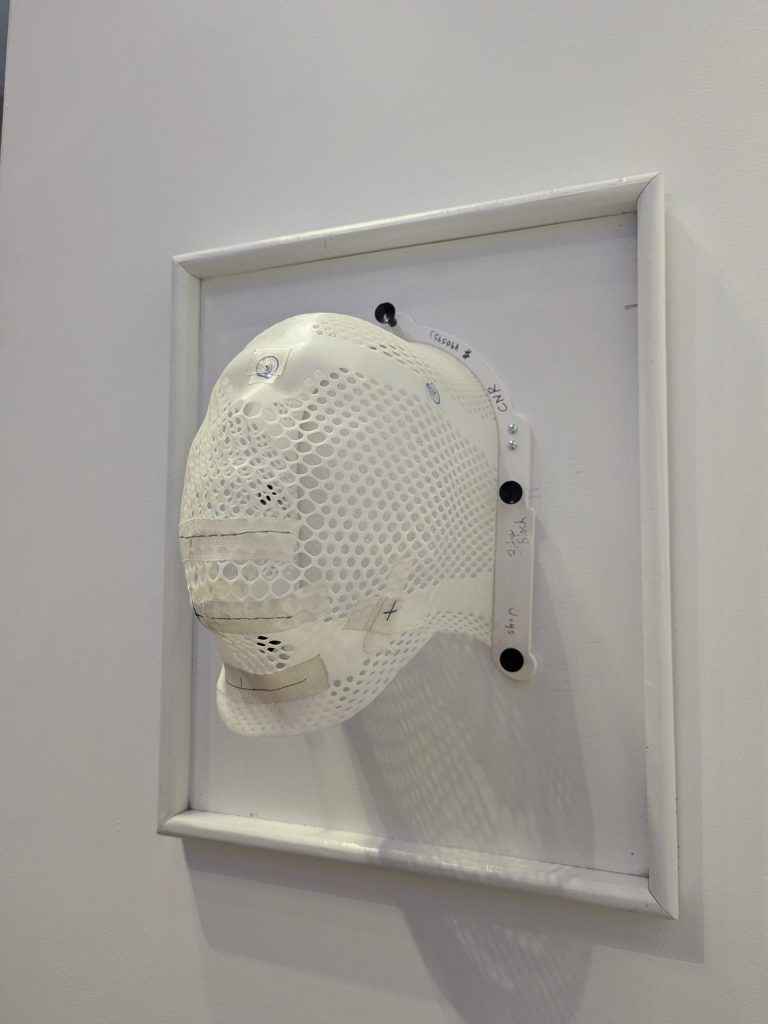A perforated white plastic mask, molded to fit a human face, is mounted on a white board within a white frame, displayed on a wall. The mask has handwritten markings and labels in pen and tape, suggesting medical or technical use.