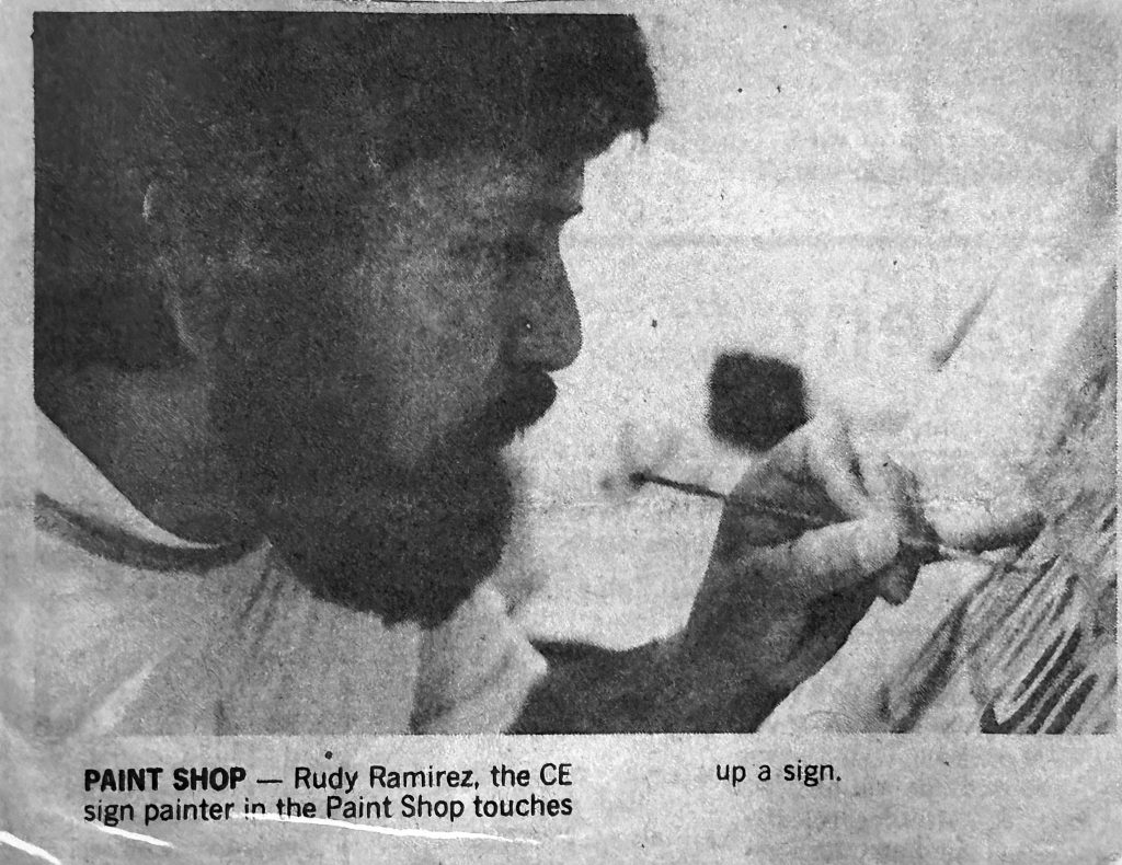 Black-and-white photo of a man, Rudy Ramirez, carefully painting a sign by hand with a small brush. His face is focused in profile as he works in close detail.