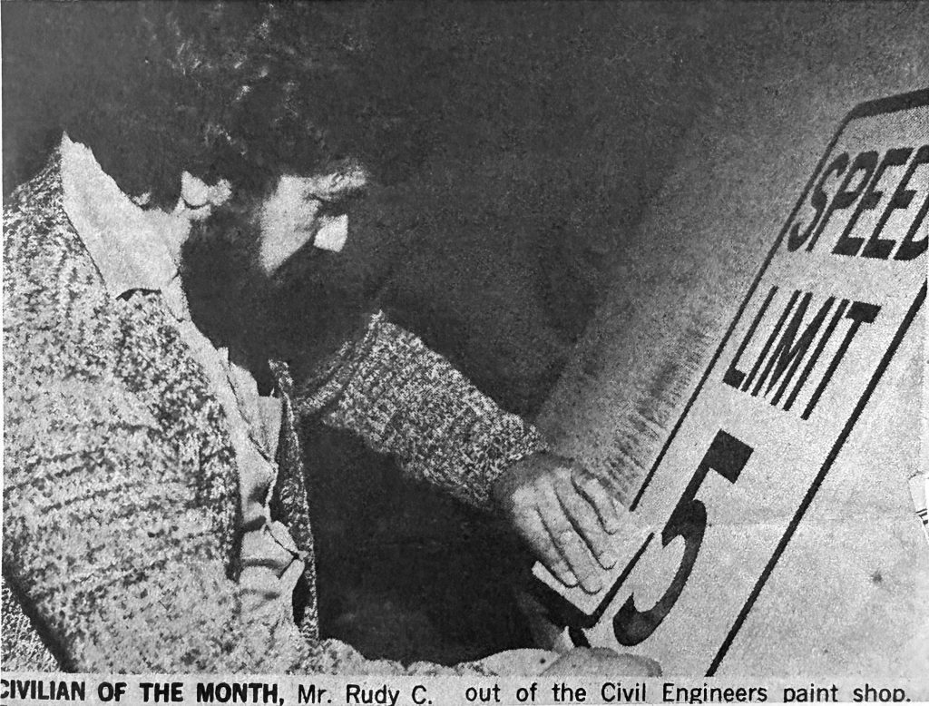 Black-and-white photo of Rudy C. Ramirez positioning or painting a large “Speed Limit 5” sign while wearing a textured cardigan. The caption notes he was named Civilian of the Month.