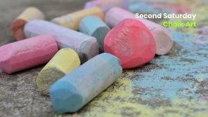 Close-up of colorful sidewalk chalk pieces scattered on pavement with text promoting a “Second Saturday” chalk art event.