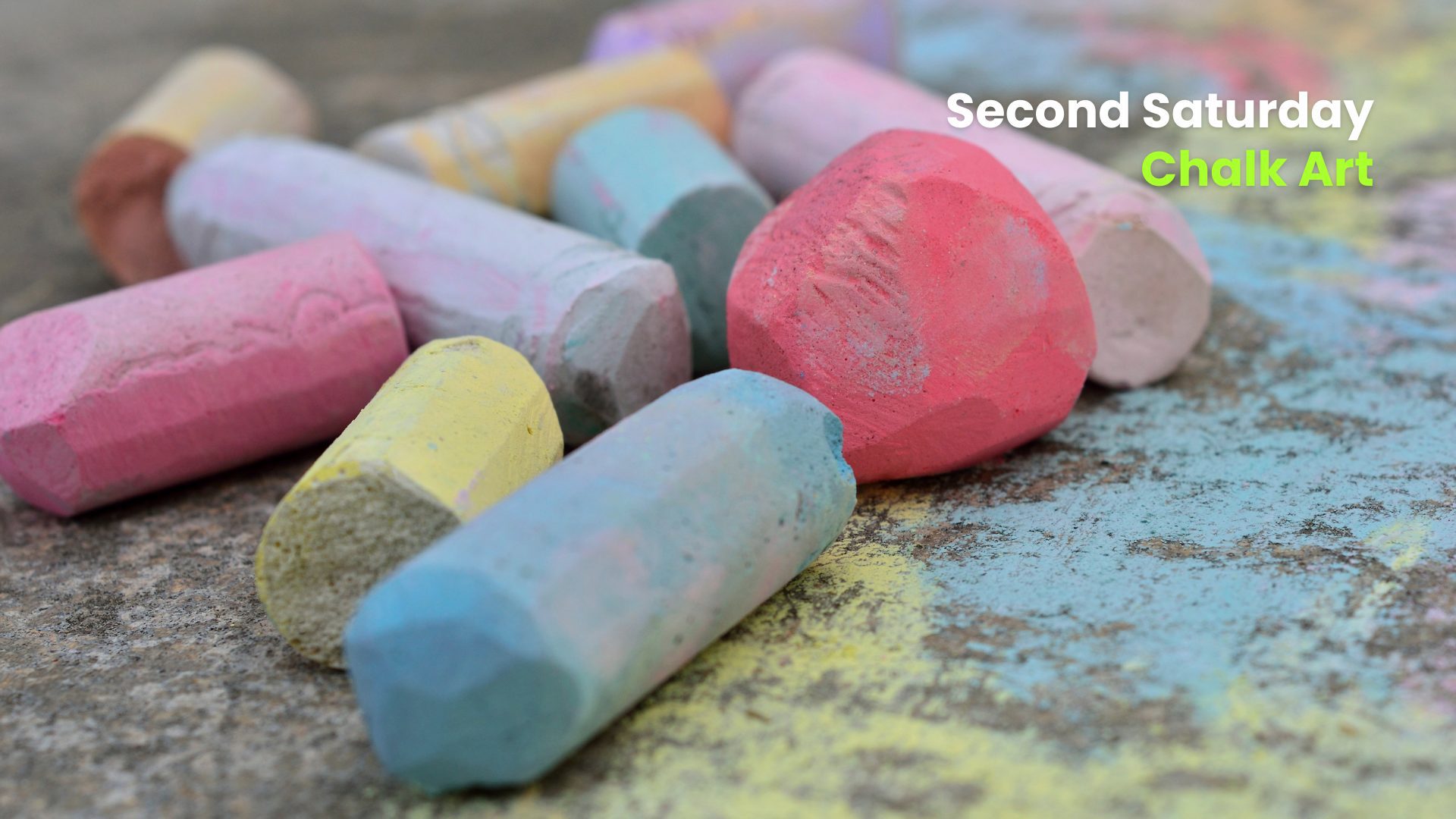 Close-up of colorful sidewalk chalk pieces scattered on pavement with text promoting a “Second Saturday” chalk art event.