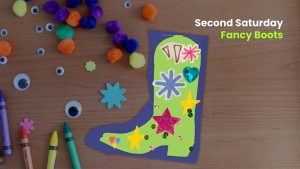 Craft table with colorful pom-poms, googly eyes, and crayons surrounding a decorated paper boot; text reads “Second Saturday Fancy Boots” in the corner.