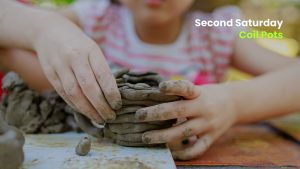 Child with clay-covered hands building a coil pot at a table during a hands-on “Second Saturday” pottery activity.