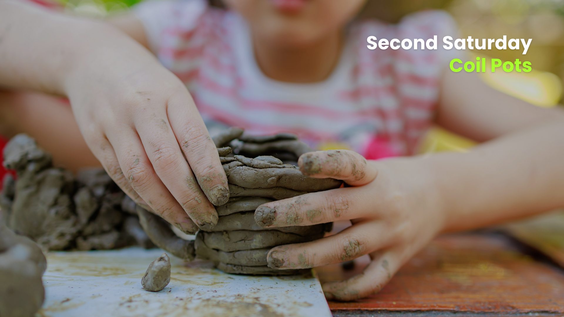 Child with clay-covered hands building a coil pot at a table during a hands-on “Second Saturday” pottery activity.
